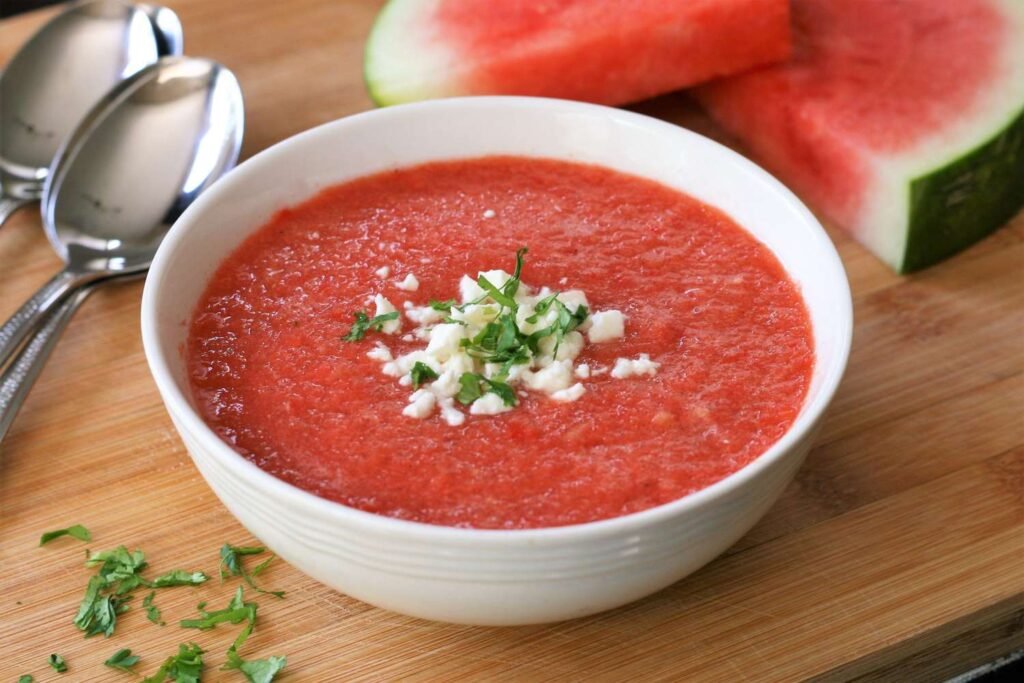 A top-down shot of a white bowl containing vibrant pink watermelon gazpacho sprinkled with chopped herbs, served on a cloth napkin.