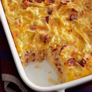 A creamy, golden-brown casserole in a white baking dish with a corner removed, revealing a moist, cheesy interior studded with crispy bacon and hash browns
