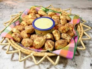 A napkin-lined basket heaped with round cinnamon roll bites dusted in cinnamon sugar, centered around a small bowl of dipping sauce—warm, inviting, and ready to enjoy