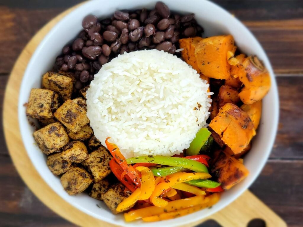 Overhead view of a grain bowl with a base of rice, topped with neatly arranged roasted sweet potato cubes, black beans, strips of colorful bell peppers, and a mound of rice or quinoa at the center.