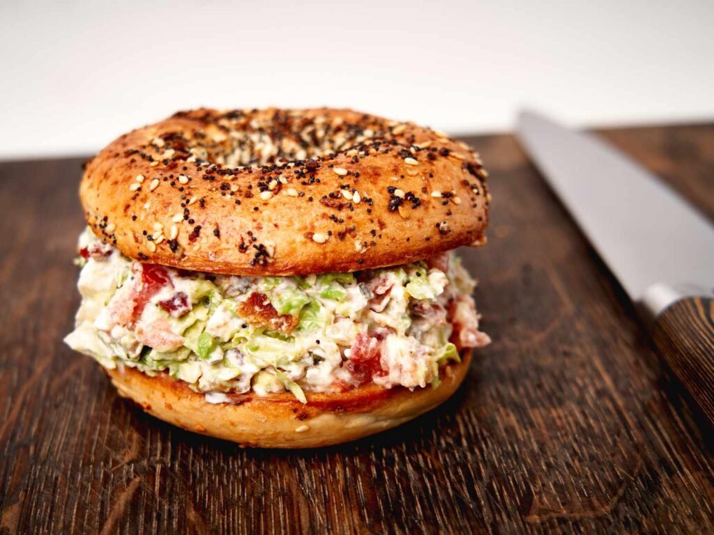 A toasted everything bagel generously filled with chopped cream cheese, crisp bacon bits, diced tomato, chopped romaine, and avocado, giving a colorful, chunky sandwich appearance.