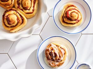 A crisp overhead shot of several spiral rolls baked to golden perfection, each featuring specks of bacon within the swirls and lightly drizzled with icing—presented on a white hexagon-tiled surface near matching dishware.
