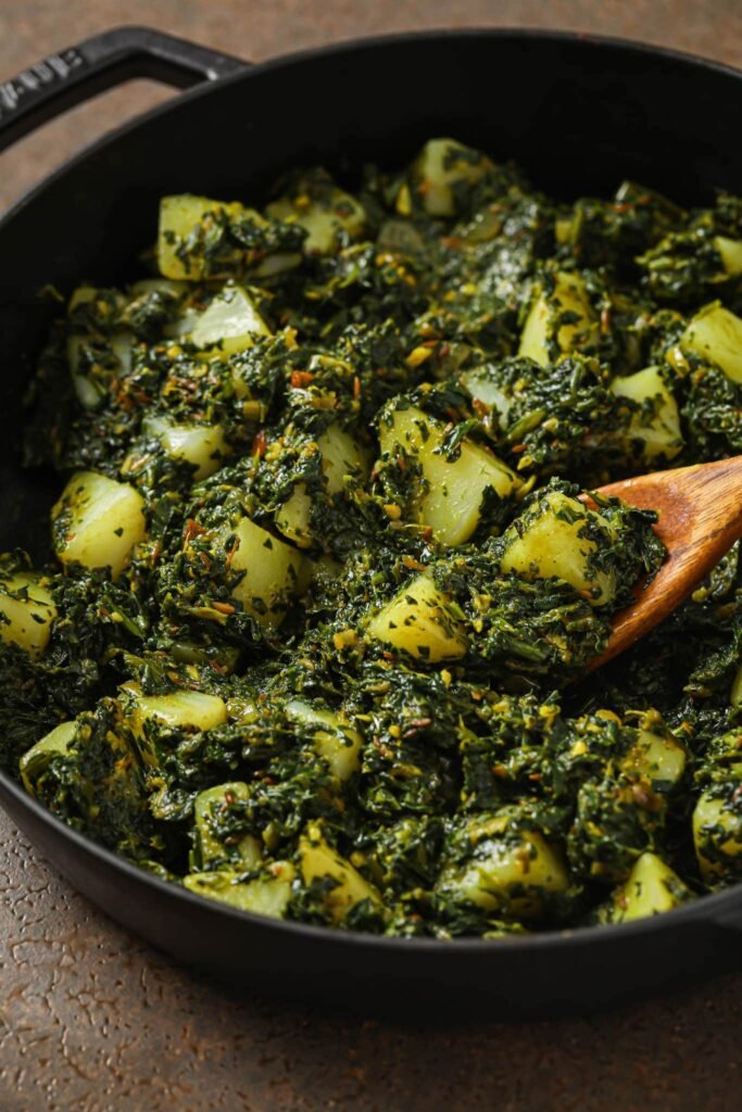 Sautéed spinach and potato curry in a dark bowl, with tender potato cubes and wilted green spinach leaves.”