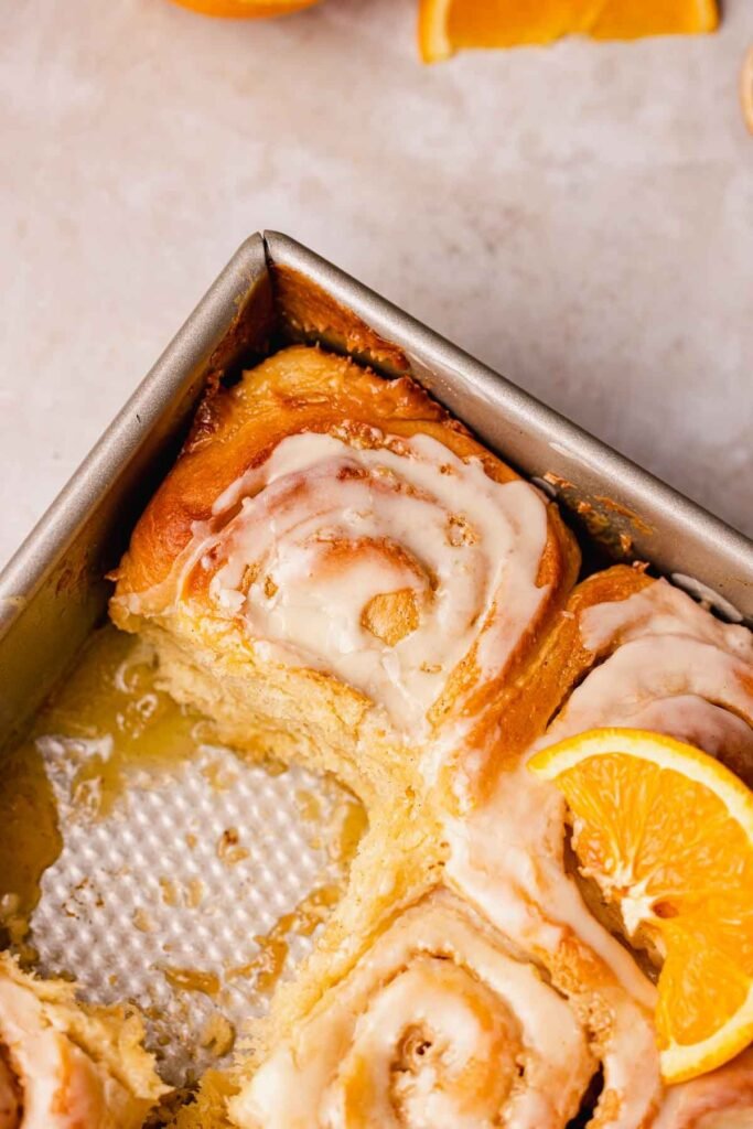“A close-up of freshly baked orange‑cinnamon rolls arranged in a baking dish, each topped with a glossy orange glaze that drizzles down the swirled layers.”