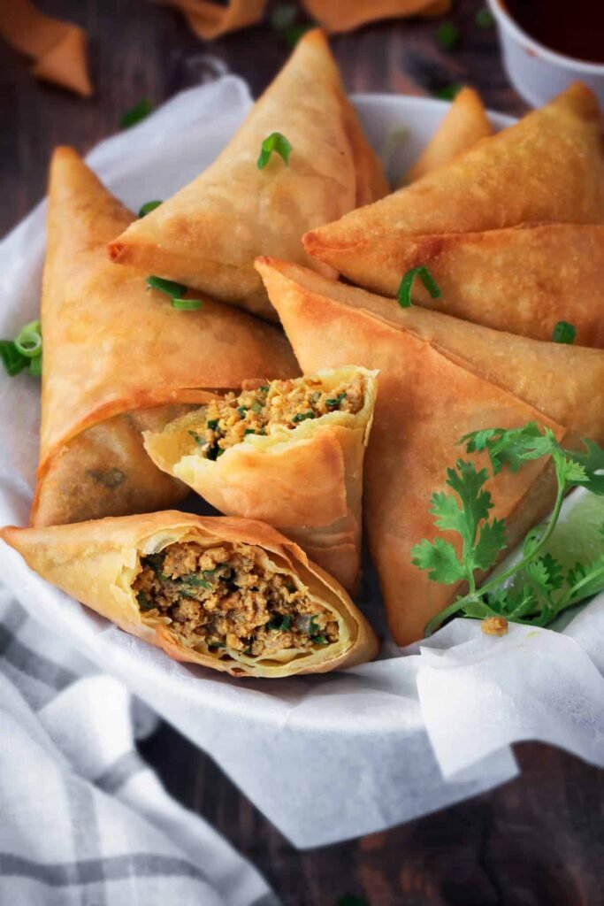 A close-up of several golden-brown triangular keema samosas arranged on a plate, with a small bowl of vibrant red dipping sauce beside them.