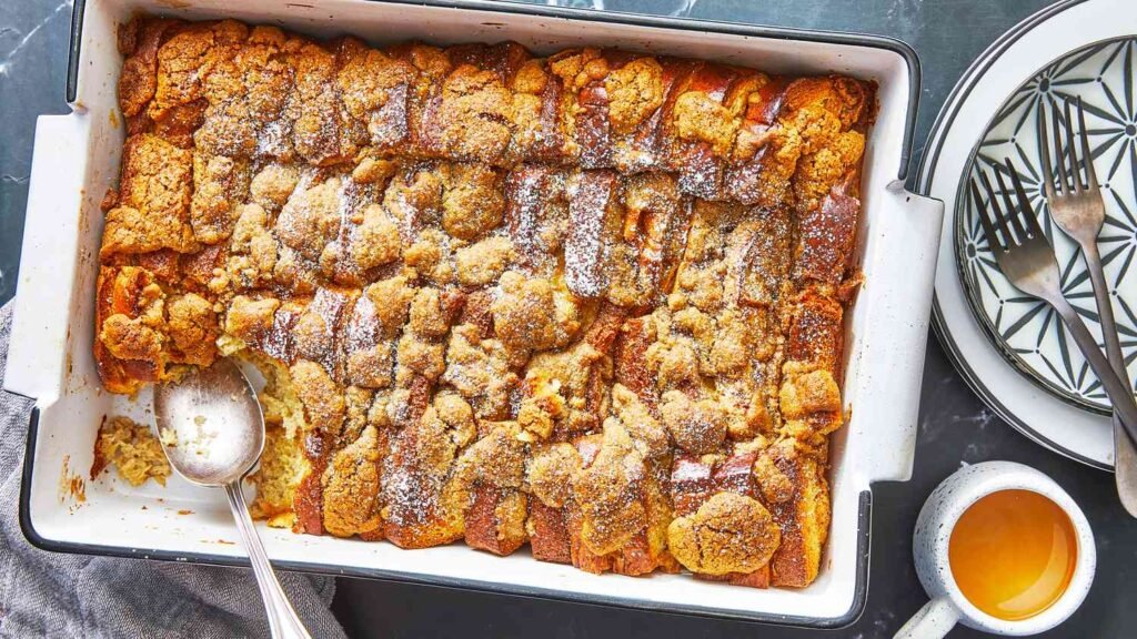Golden-brown brioche French toast casserole in a white rectangular baking dish with a lightly browned streusel topping, served next to a mug of coffee