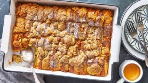 Golden-brown brioche French toast casserole in a white rectangular baking dish with a lightly browned streusel topping, served next to a mug of coffee