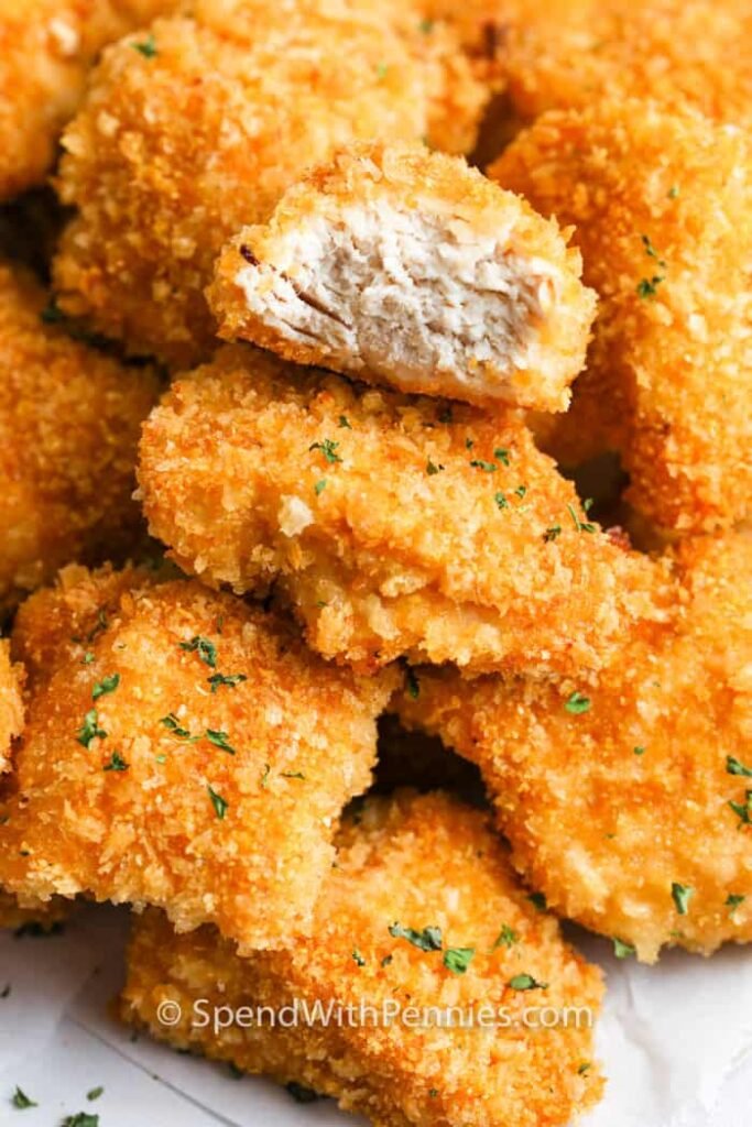 Heap of oven‑baked chicken nuggets on a tray—each nugget has a flaky, golden crust; background shows a warm kitchen setting hinting at freshness.
