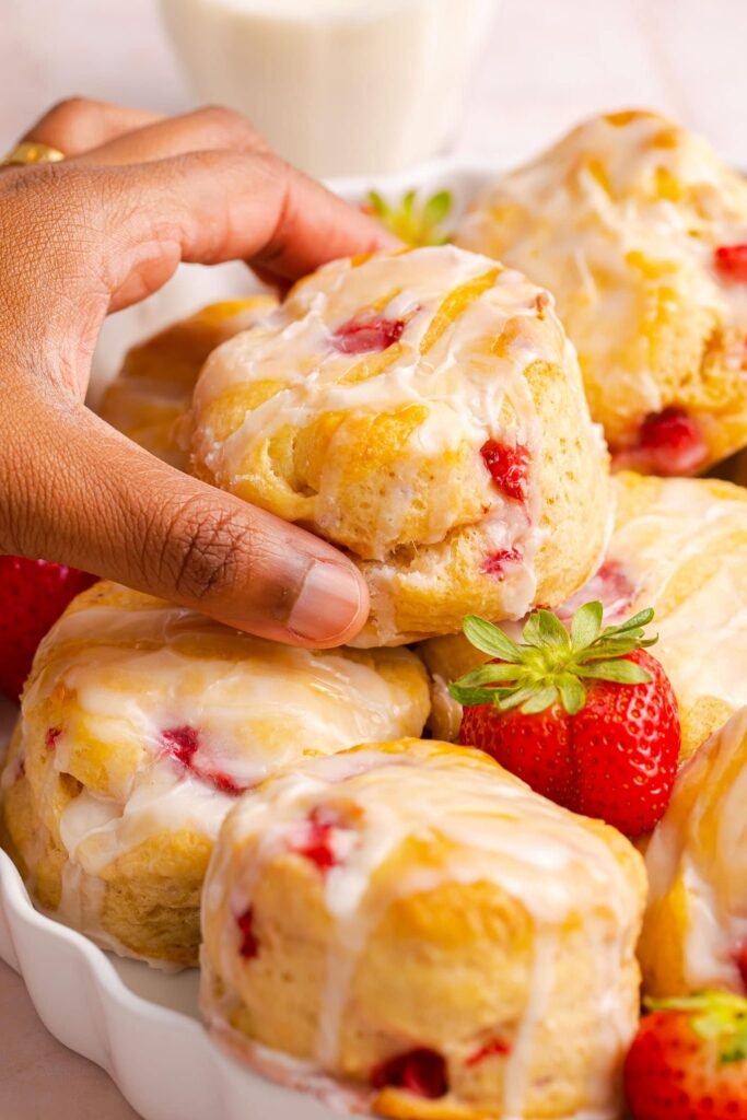 “Hand grabbing a glazed strawberry biscuit dotted with fresh strawberry pieces.”