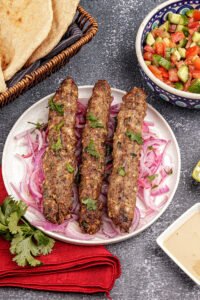 Four cylindrical grilled beef seekh kebabs on metal skewers, placed on a round white plate with scattered red chili flakes and fresh parsley.