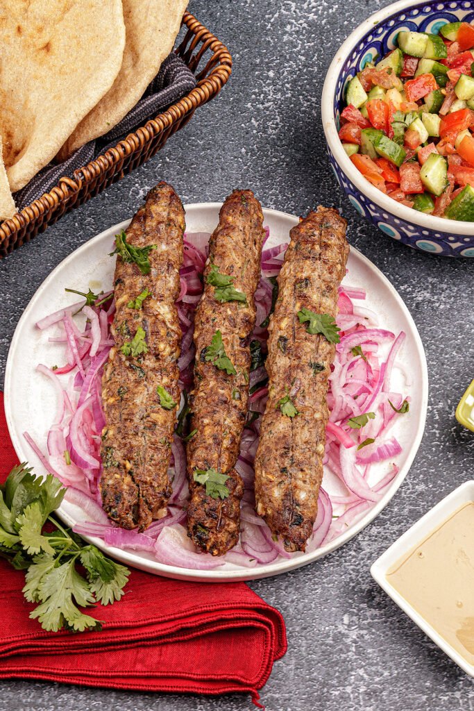 Four cylindrical grilled beef seekh kebabs on metal skewers, placed on a round white plate with scattered red chili flakes and fresh parsley.