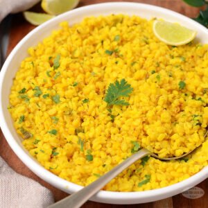 A white shallow bowl filled with vibrant yellow dry moong dal, with individual lentils lightly separated. A silver spoon rests in the dish, and a garnish of fresh green coriander adds a pop of color.