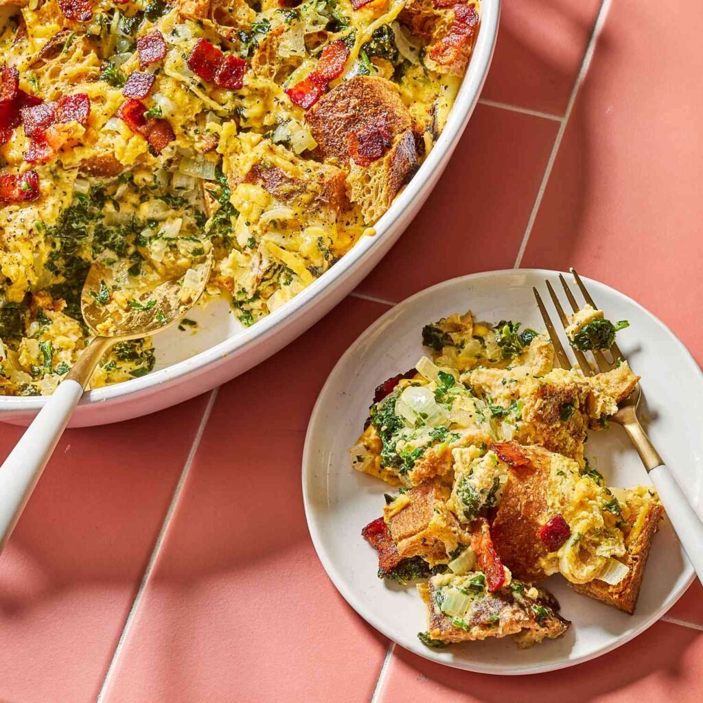 : A white oval baking dish filled with golden-brown strata—a baked casserole of cubed bread, bits of crispy bacon, spinach, and melted cheddar cheese—served with a spoon and small plate.