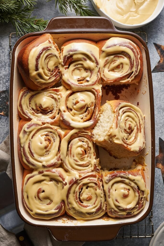 : Overhead view of eight soft cinnamon rolls in a baking dish, topped with swirls of rich frosting and dusted with holiday sprinkles, suggesting a Christmas morning treat.