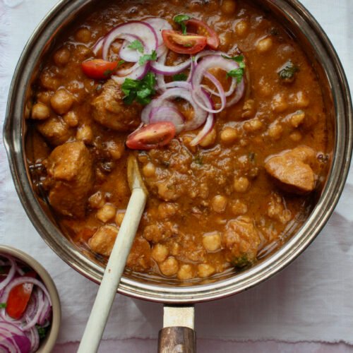 A top-down view of a pot filled with rich, reddish-brown curry featuring tender chicken pieces and plump chickpeas, garnished with thinly sliced red onions and fresh cilantro—evoking hearty, home-style cooking.