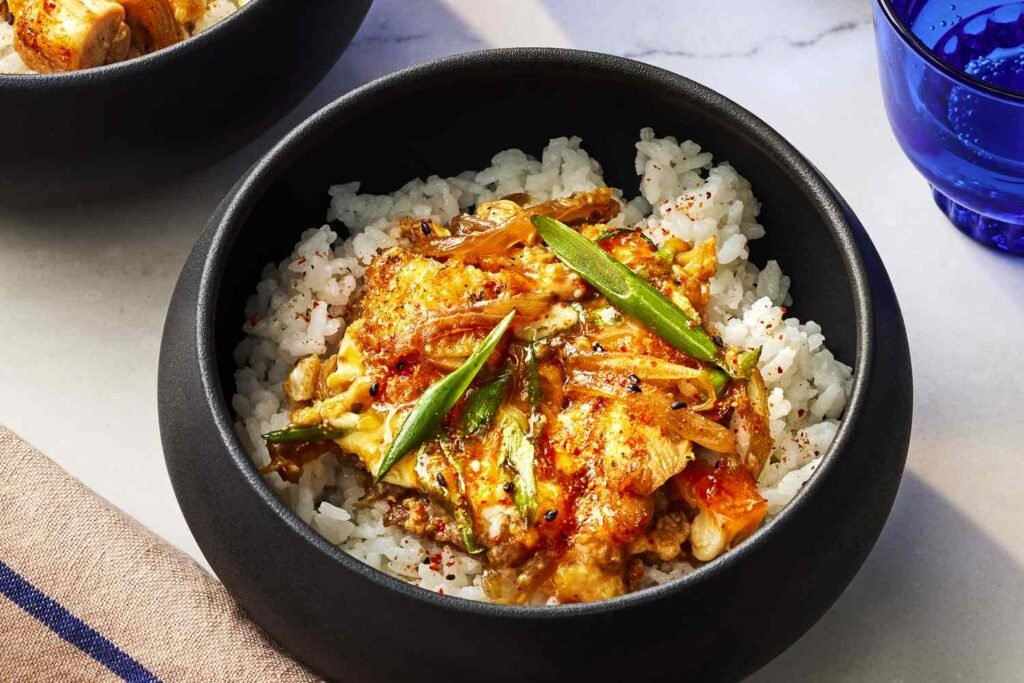 A black donburi bowl filled with steamed rice, tender chicken pieces and onions simmered in golden dashi‑soy broth, topped with gently set egg and scallions.