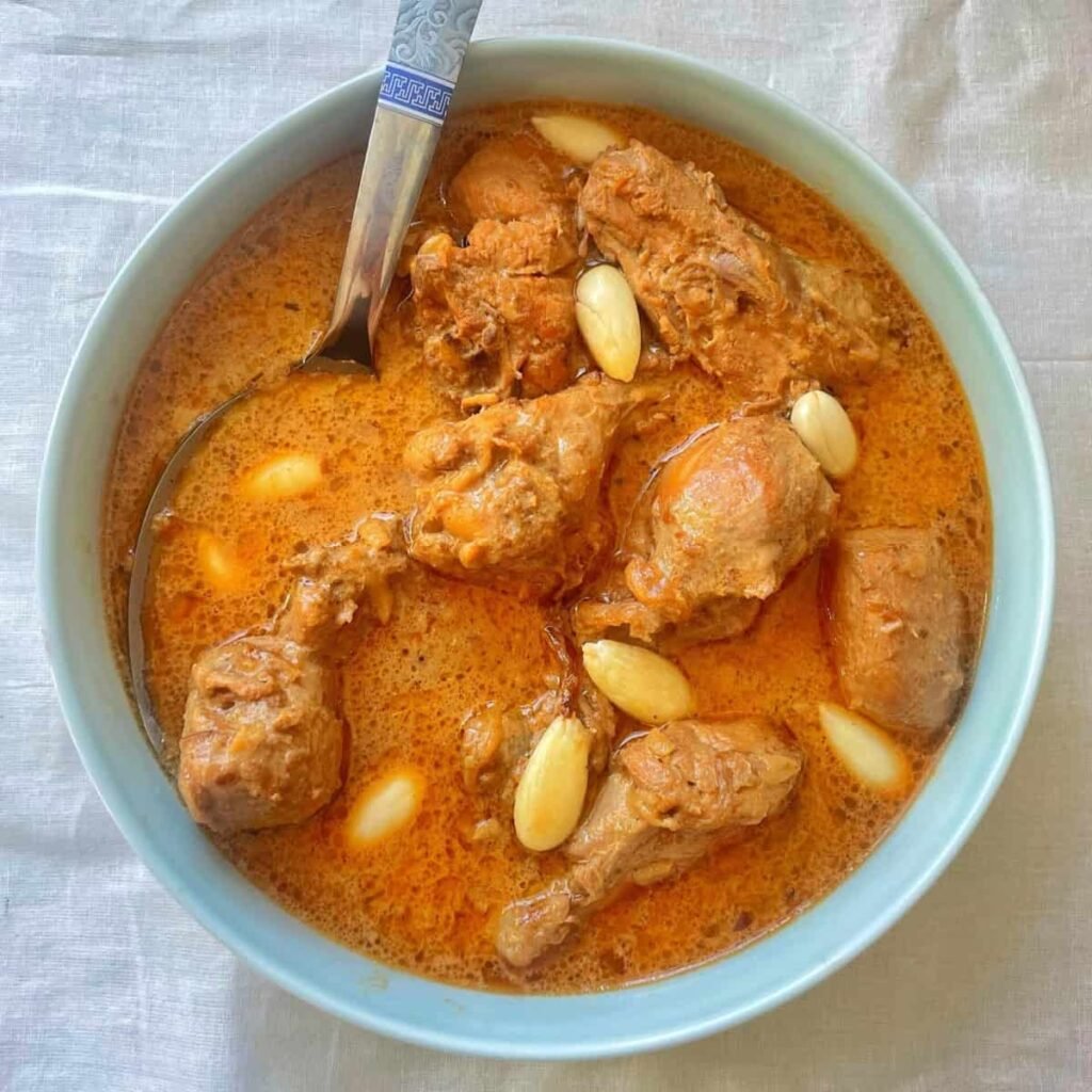 Top-down shot of a bowl of chicken qorma in a light orange-yogurt base, dotted with slivered almonds and tender chicken pieces, showcasing its creamy texture.