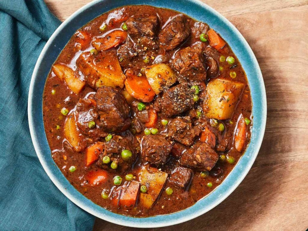Overhead view of a blue bowl filled with chunky beef stew—tender beef, carrots, potatoes, and peas in a thick brown gravy.”