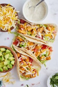 Three soft flour tortillas on a white plate, filled with shredded ranch‑seasoned chicken, topped with diced avocado and a drizzle of creamy ranch sauce, set beside a small bowl of avocado.
