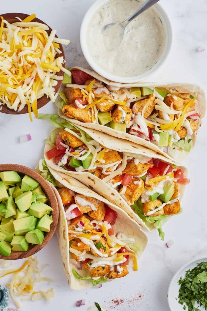 Three soft flour tortillas on a white plate, filled with shredded ranch‑seasoned chicken, topped with diced avocado and a drizzle of creamy ranch sauce, set beside a small bowl of avocado.