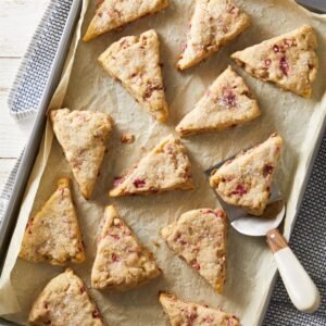 “Triangular rhubarb scones on parchment: golden-brown and flaky with visible pink rhubarb chunks, some glazed lightly, showing crisp edges and a tender interior.”