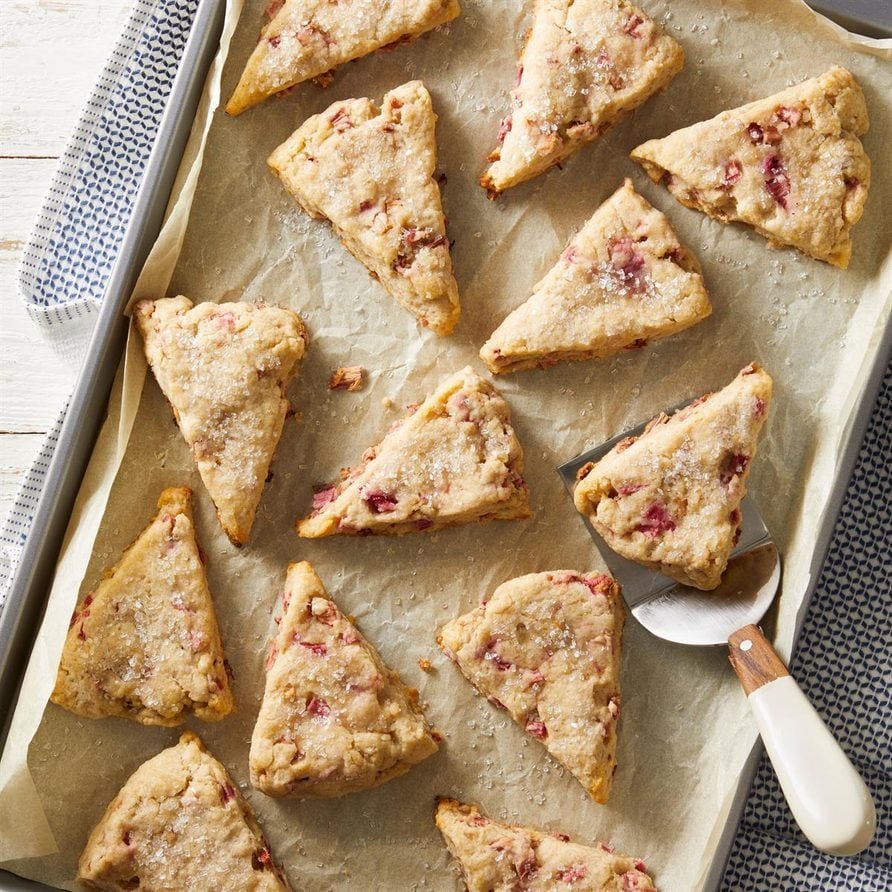 “Triangular rhubarb scones on parchment: golden-brown and flaky with visible pink rhubarb chunks, some glazed lightly, showing crisp edges and a tender interior.”