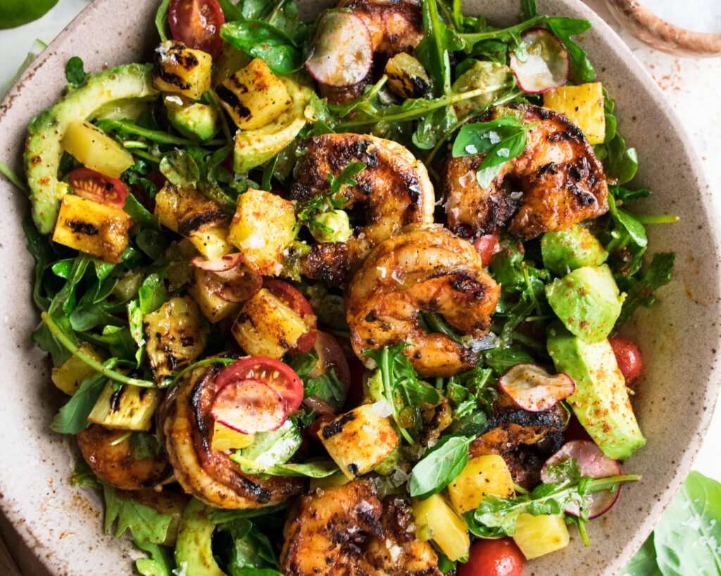 A top-down view of a summery bowl filled with grilled shrimp, vibrant mixed greens, diced avocado, cherry tomatoes, sweet corn kernels, and cucumber, all drizzled with a light vinaigrette and served in a shallow, round, light-colored bowl
