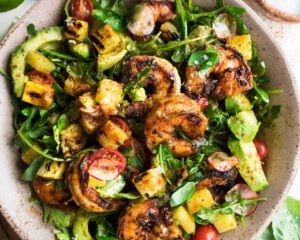 A top-down view of a summery bowl filled with grilled shrimp, vibrant mixed greens, diced avocado, cherry tomatoes, sweet corn kernels, and cucumber, all drizzled with a light vinaigrette and served in a shallow, round, light-colored bowl
