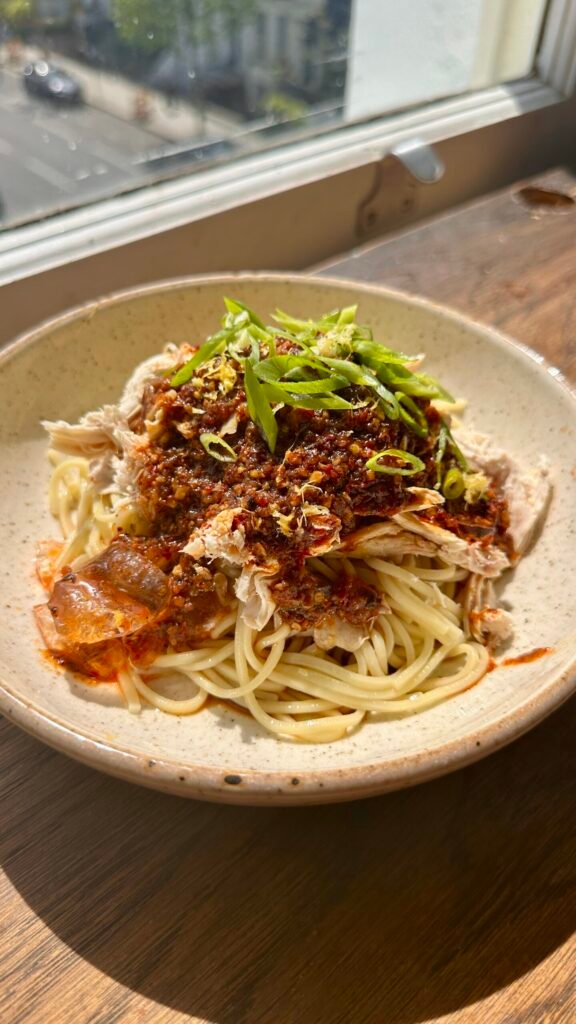 A bowl of Sichuan-style spicy chicken noodles—noodles tossed in richly colored chili oil, topped with tender shredded chicken and fresh green onion.