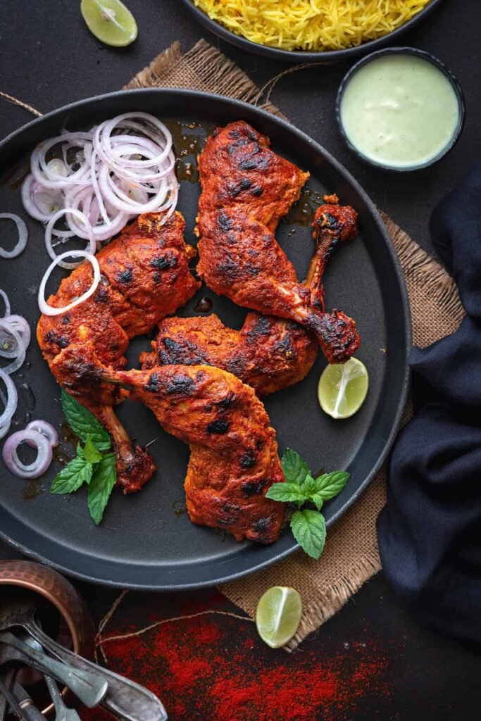 Charred, vibrant-red half‑chicken sections on a black plate, served alongside green chutney, lime slices, and onion rings—an appetizing restaurant-style presentation.