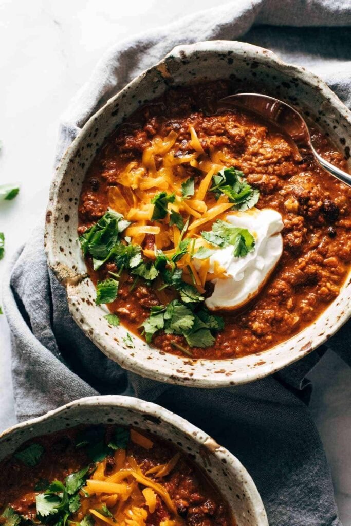 White bowl of thick vegetarian chili with beans and tomatoes, garnished with fresh cilantro—served next to lime wedges.”