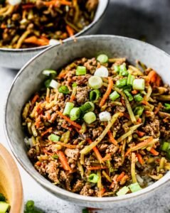 A gray bowl filled with ground meat and shredded cabbage stir-fried together, topped with chopped green onions; a second bowl sits in the background.