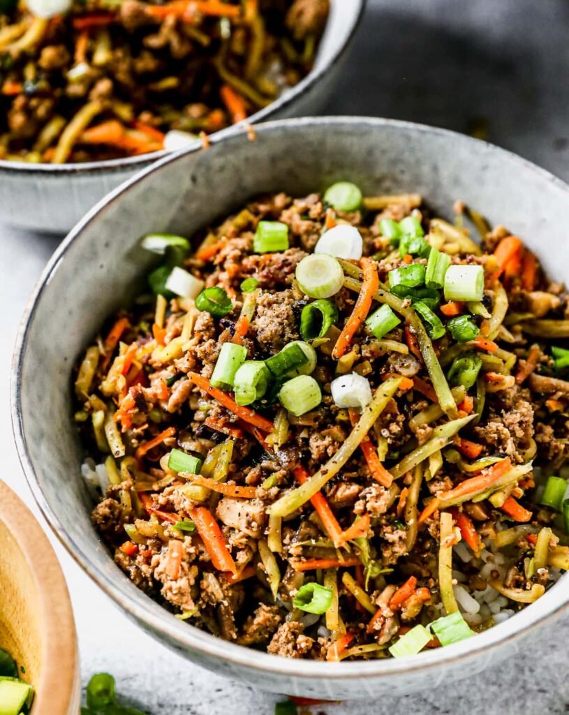 A gray bowl filled with ground meat and shredded cabbage stir-fried together, topped with chopped green onions; a second bowl sits in the background.