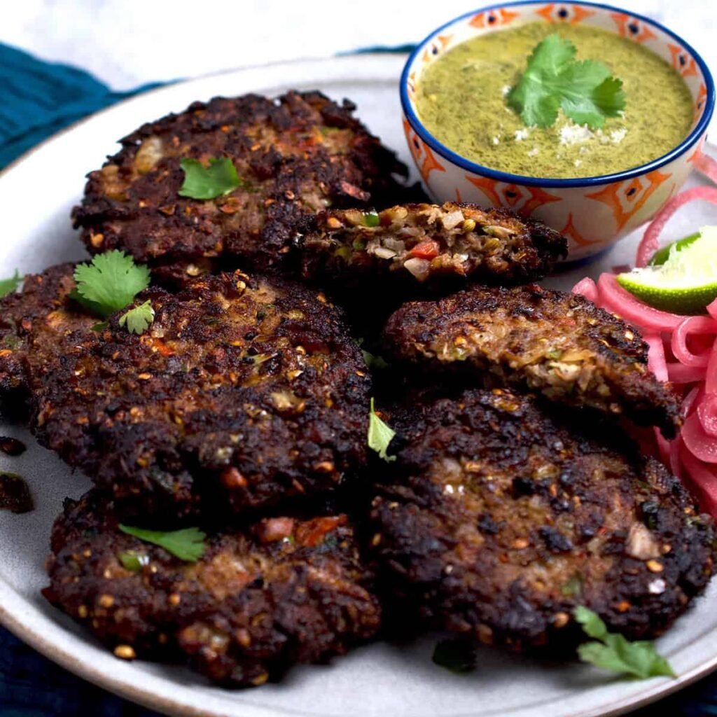 “Several golden-brown chapli kababs arranged on a plate with a small bowl of green chutney on the side.”