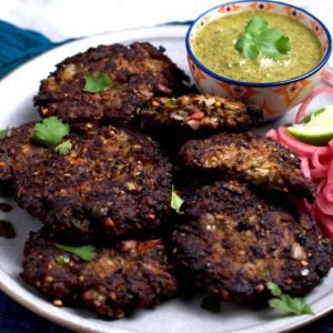  “Several golden-brown chapli kababs arranged on a plate with a small bowl of green chutney on the side.”
