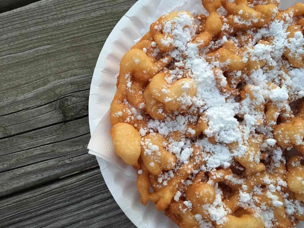 “Golden-brown spiral funnel cake dusted with powdered sugar on a white plate.”