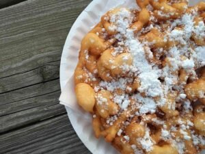 “Golden-brown spiral funnel cake dusted with powdered sugar on a white plate.”