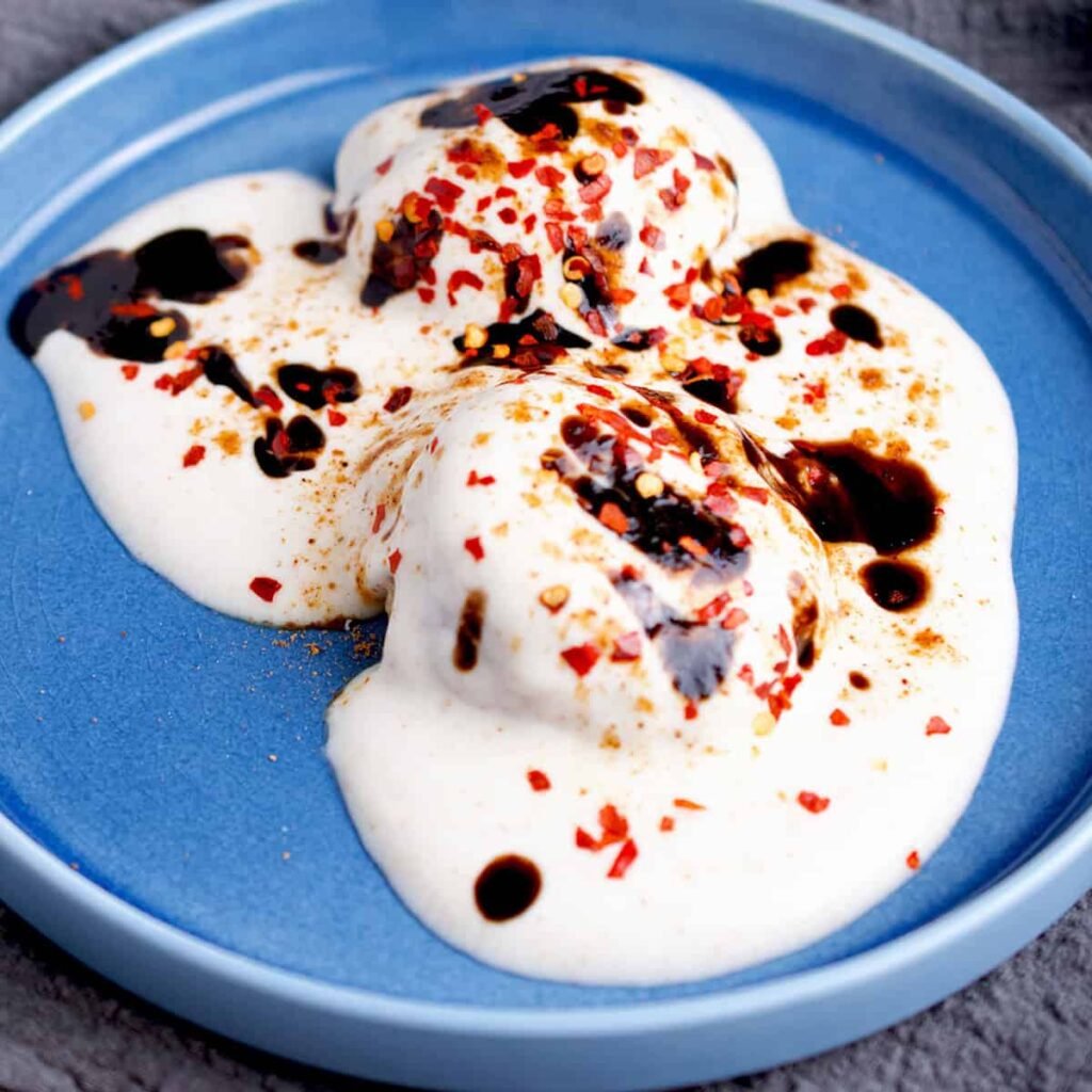 A trio of soft, yogurt-drenched lentil fritters on a blue plate, topped with dark tamarind chutney and sprinkled with red chili flakes