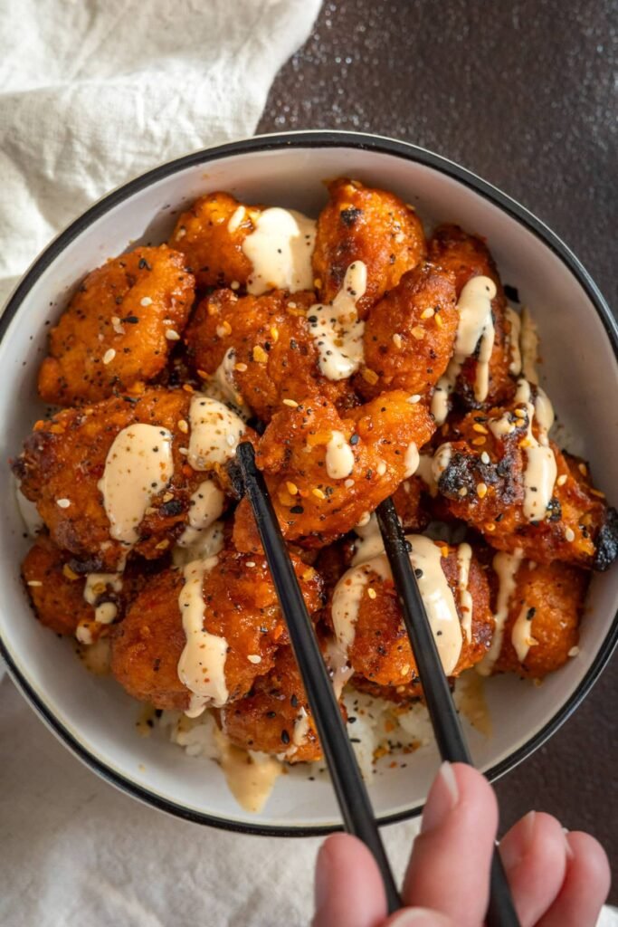 Bowl of crispy chicken chunks coated in bright orange, creamy dynamite sauce, garnished with chopped scallions