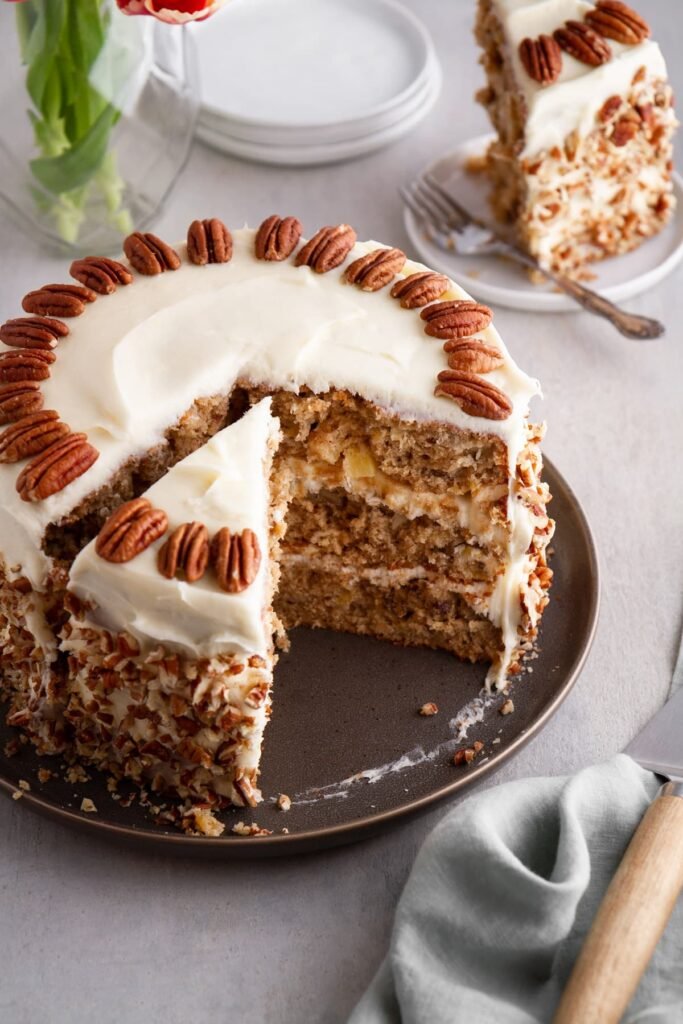 “A three-layer Hummingbird Cake with visible layers of banana‑pineapple‑pecan sponge, generously frosted with creamy white cream cheese icing. The sides are decorated using a petal or ruffle piping technique, and the cake is presented on a glass cake stand against a teal background.”
