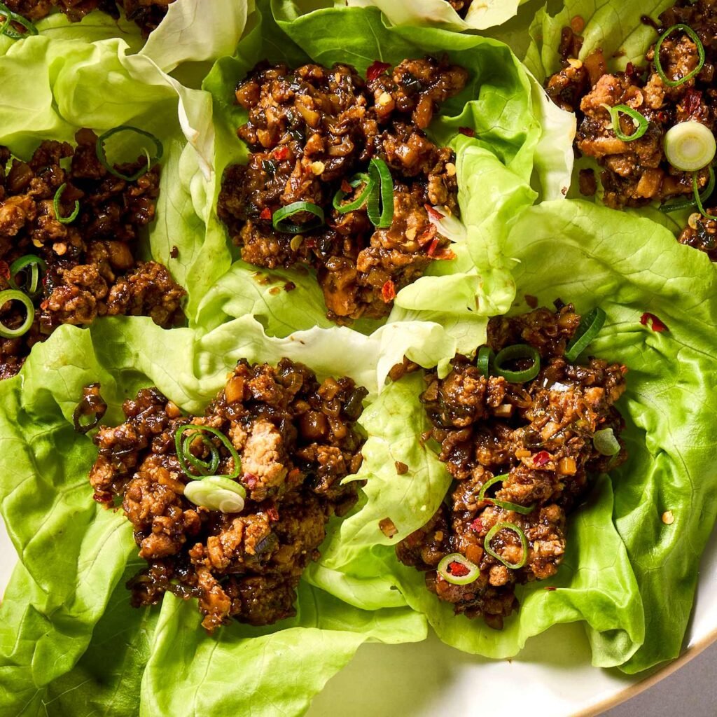 “Buttery lettuce cups filled with seasoned ground meat and vegetables, topped with chopped peanuts and scallions, served on a white plate.”