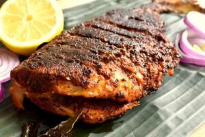 A pan‑fried whole fish coated in spicy masala, with a crisp darkened crust, served alongside wedges of lemon and garnished with onion rings.