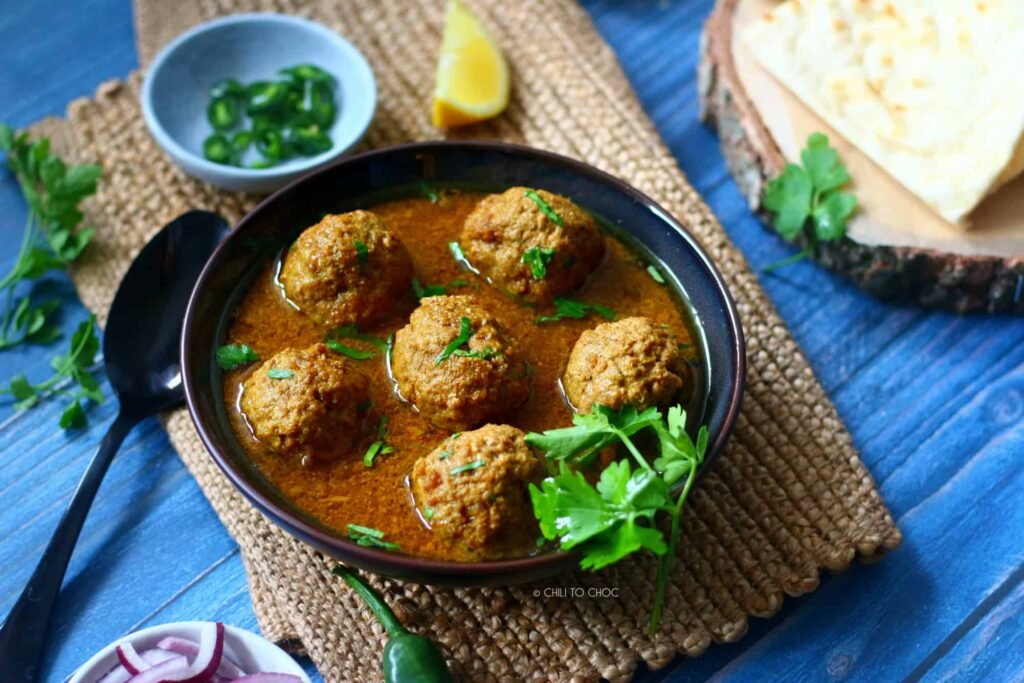A black skillet brimming with beef koftas nestled in a thick, spicy curry, garnished with scattered fresh cilantro leaves and green chillies in the background.