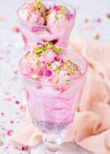  Two glasses of falooda featuring vivid layers of rose syrup, basil seeds, noodles, milk, garnished with rose petals and chopped nuts; spoons inserted—captures a playful, festive feel.