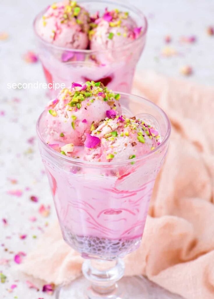 Two glasses of falooda featuring vivid layers of rose syrup, basil seeds, noodles, milk, garnished with rose petals and chopped nuts; spoons inserted—captures a playful, festive feel.