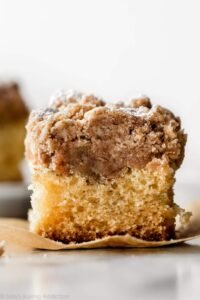 “Squared slice of New York‑style crumb cake on a plate: a tender, buttery yellow cake base topped with a thick, brown sugar‑cinnamon crumb streusel, lightly dusted with powdered sugar.”