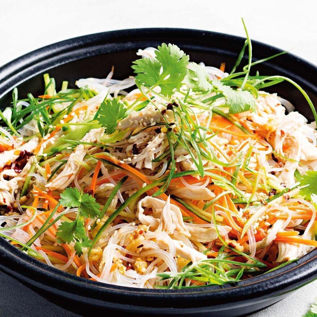 Cold chicken noodle salad in a bowl with colorful vegetables, sesame seeds, and chopsticks on the side.