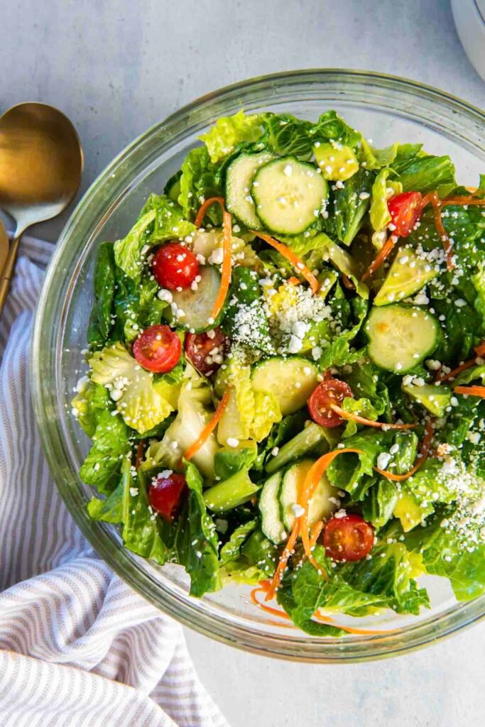 Mixed green salad with cherry tomatoes, cucumber, sunflower seeds, and wooden serving spoons in a large white bowl.