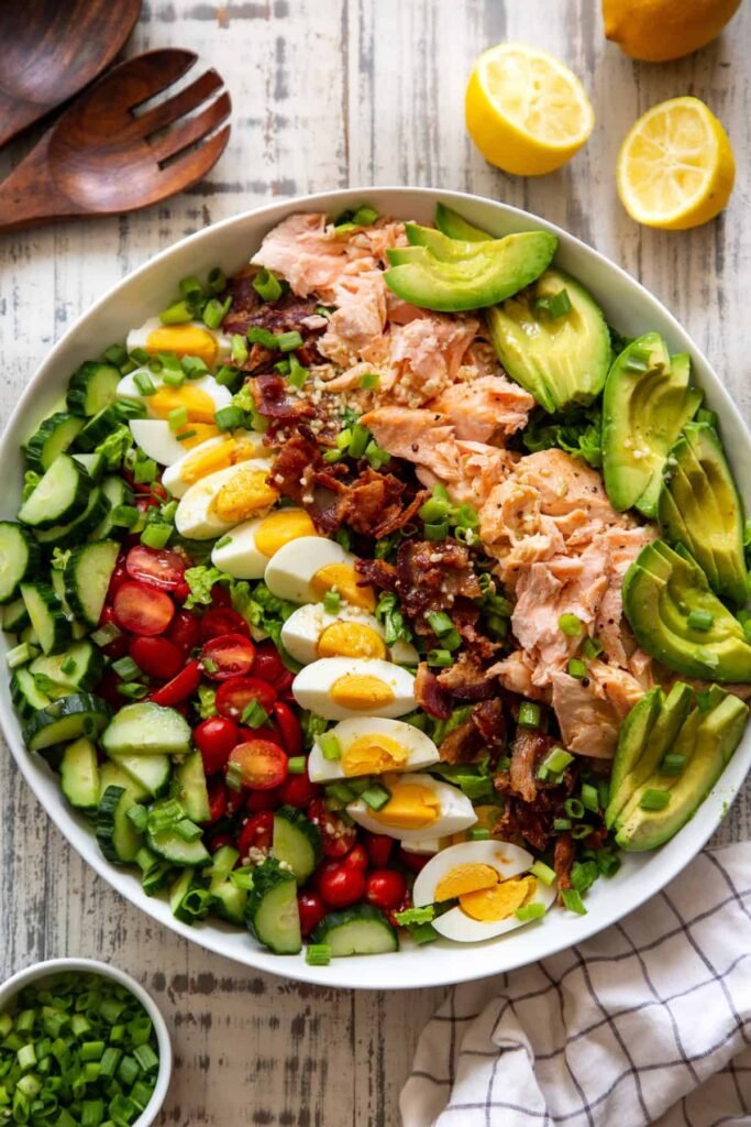 A top-down view of a composed bowl featuring flaked salmon fillets, halved hard‑boiled eggs, ripe avocado slices, cherry tomatoes, sweet corn kernels, cucumber rounds, crisp mixed greens, and crumbled bacon, all arranged in colorful rows and drizzled with a light vinaigrette