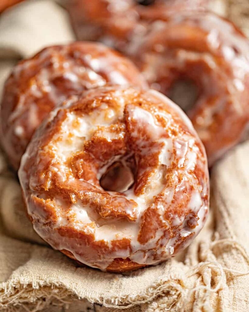 A stack of golden-brown sour cream donuts with a glossy crackled vanilla glaze, showing the soft, cakey texture beneath.”
